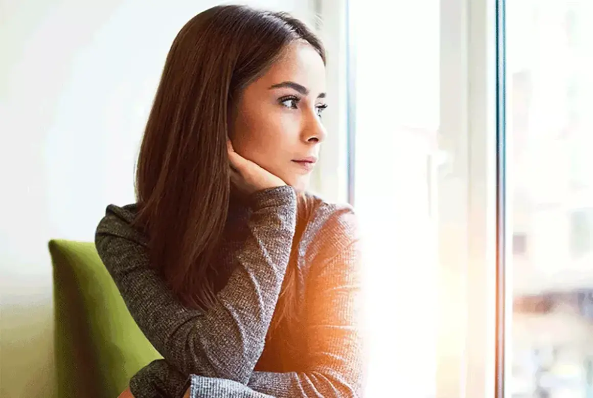Donna pensierosa guarda fuori dalla finestra, preoccupata per un ciclo in ritardo e come farlo venire.