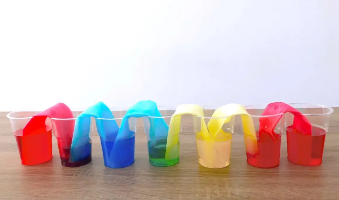 Colorati esperimenti per bambini: strisce di carta assorbono liquidi colorati in bicchieri trasparenti, creando un arcobaleno liquido.