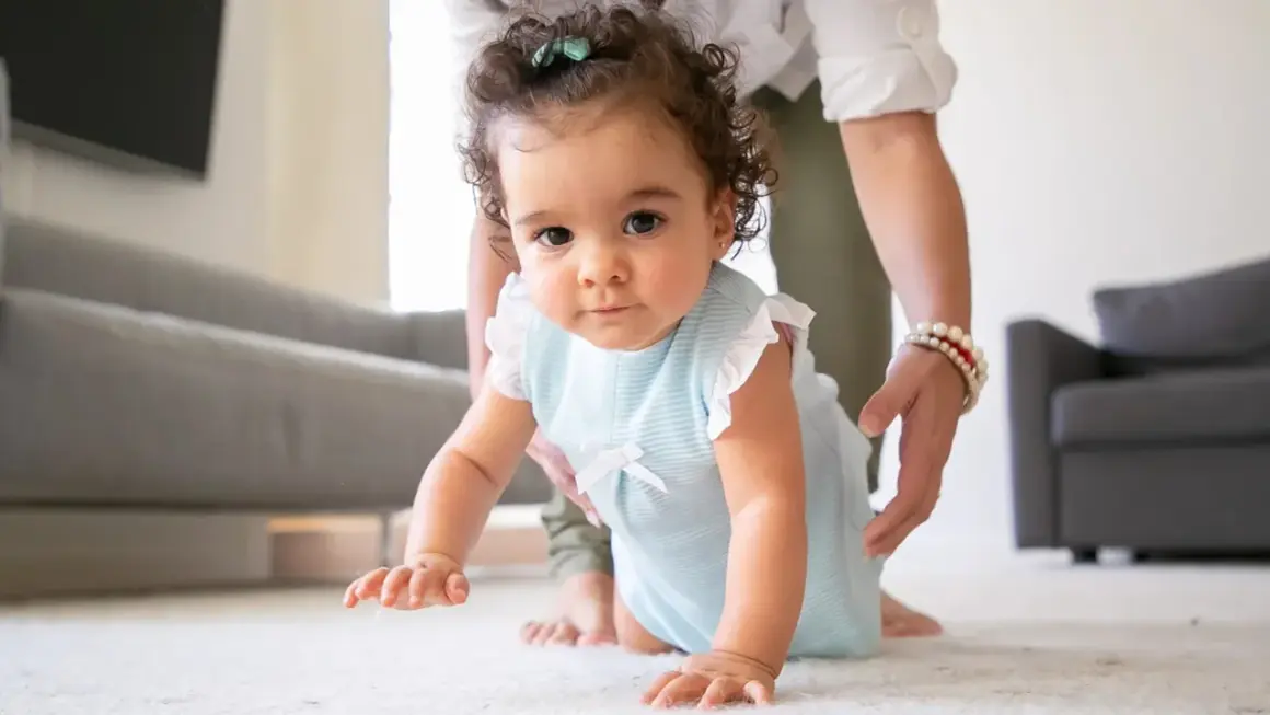 A baby in a blue striped outfit crawls on a white rug, with a person's hands gently supporting them.
