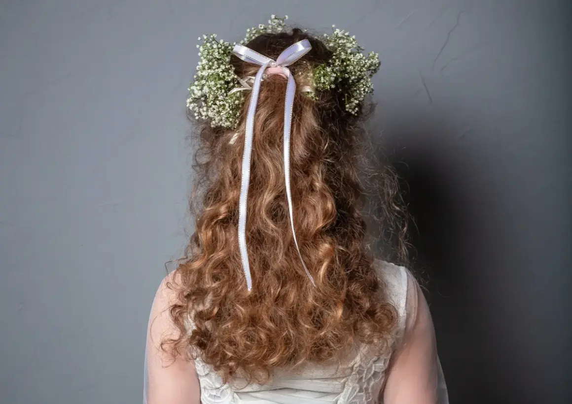 Capelli ricci con coroncina di fiori e nastri, perfetti per trecce acconciature comunione.
