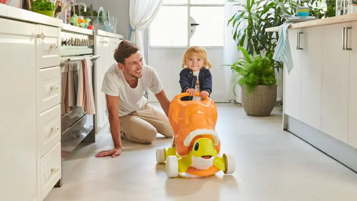 Un papà sorridente incoraggia i primi passi del suo bambino su un cavalcabile a forma di tartaruga in cucina.