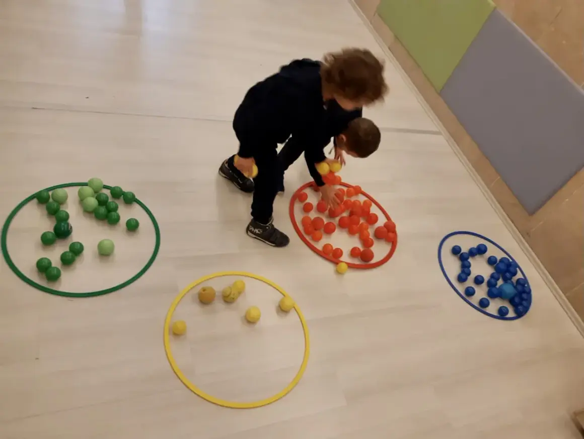 Bambini 3-5 anni impegnati in giochi motori, spostano palline colorate tra cerchi sul pavimento.