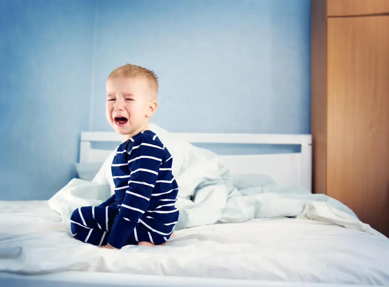 Un bambino di 2 anni si sveglia piangendo disperato sul letto, con pigiama a righe blu e bianche.