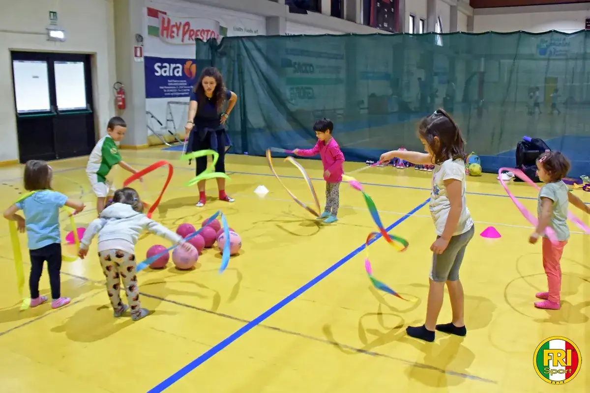 Bambini 3-5 anni si divertono con nastri colorati e palloni durante giochi motori in palestra.