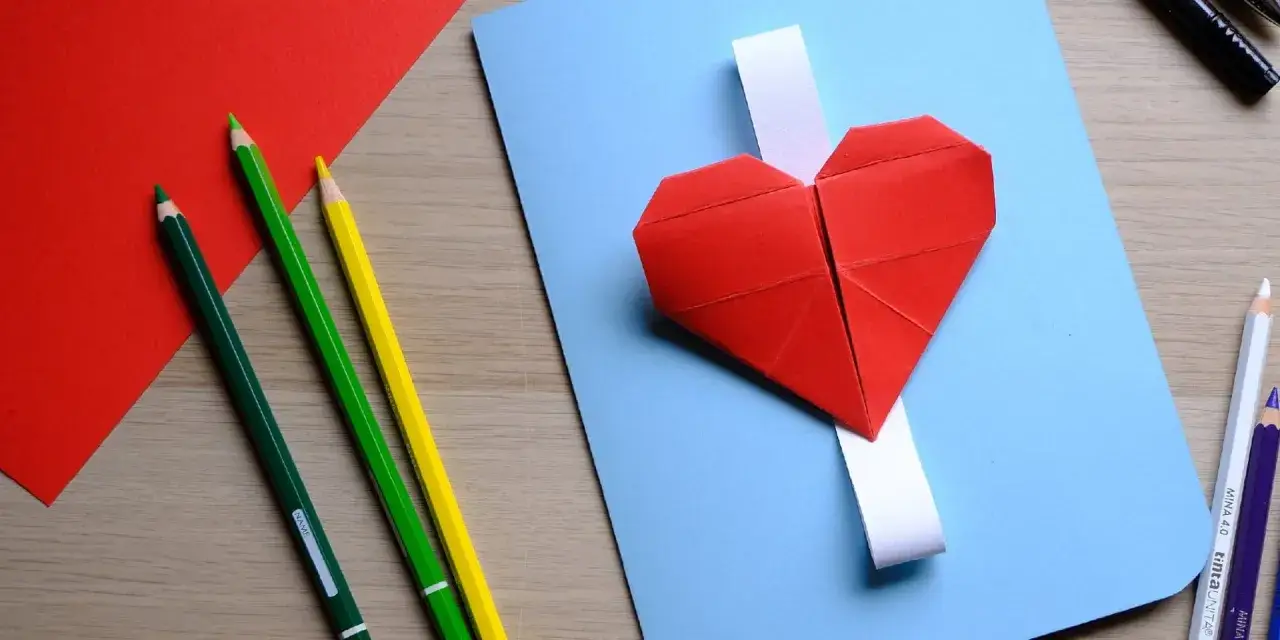 Un cuore rosso origami su un foglio azzurro, pronto per la festa della nonna. Matite colorate e fogli rossi completano la scena creativa.