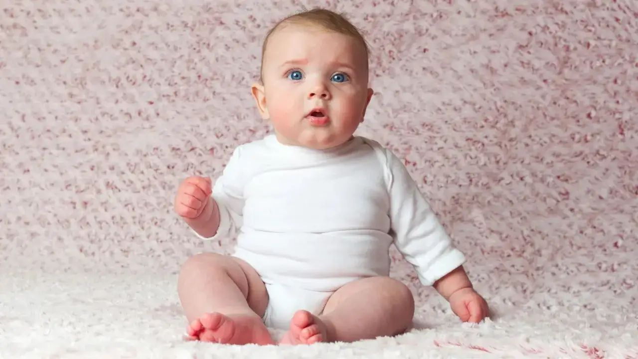 Neonato di 6 mesi con occhi azzurri, indossa una tutina bianca e si appoggia sulle mani, non sta ancora seduto da solo.