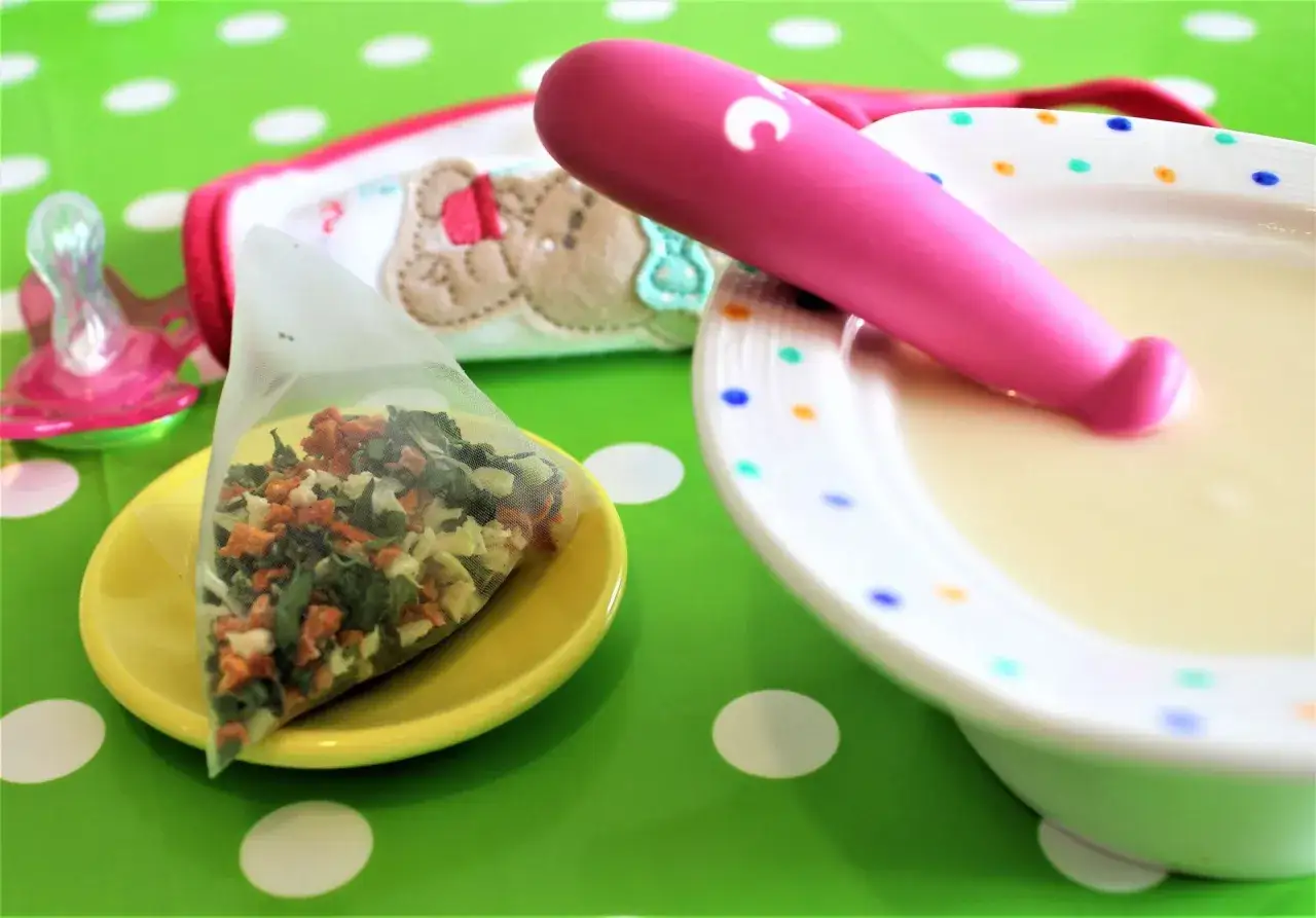 Pappa e tisana per bimbi di 2 anni: un bavaglino con orsetto, ciuccio, bustina di verdure e ciotola con pappa.