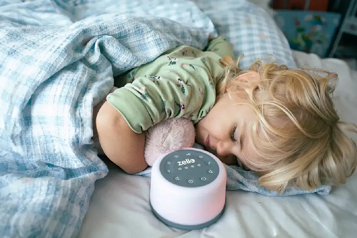 Bambino dorme sereno accanto a un dispositivo per rumori bianchi neonato, avvolto in una coperta azzurra.