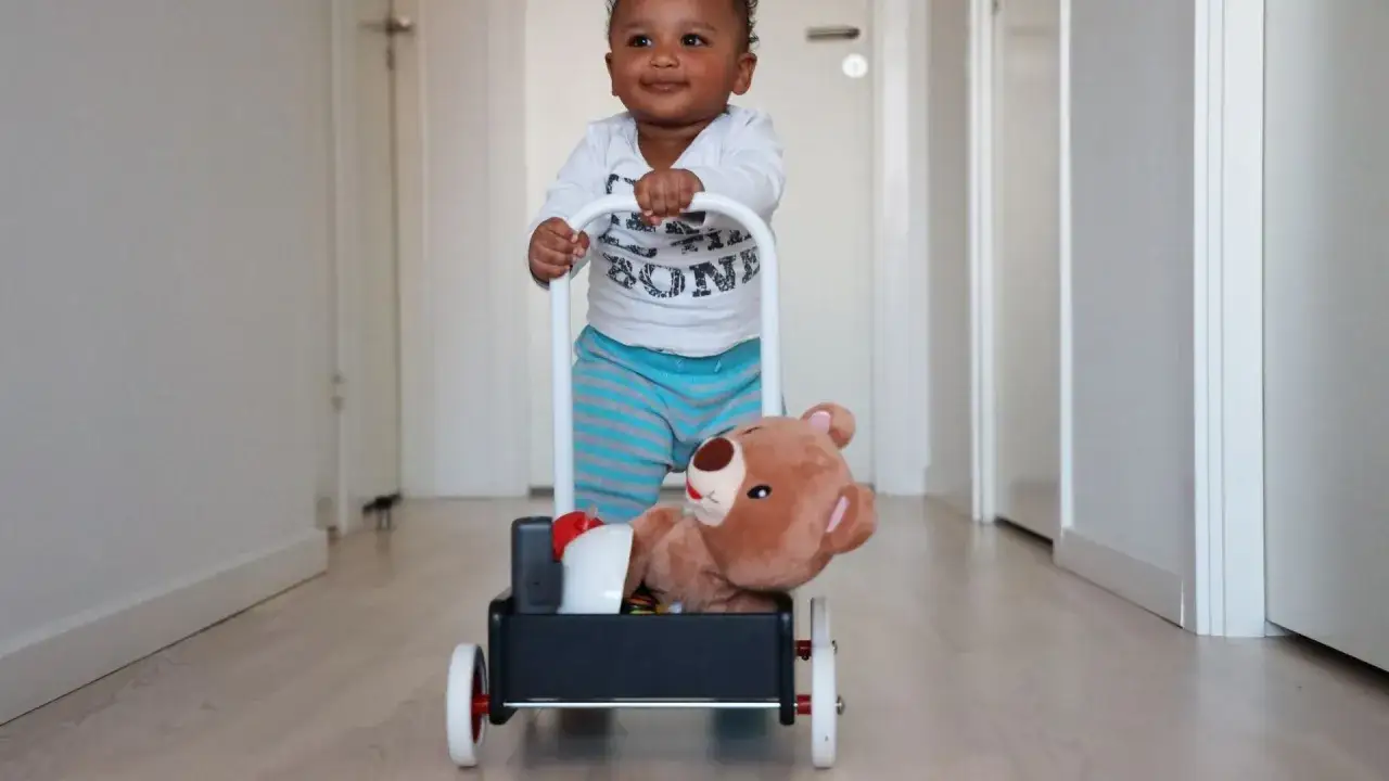 Bambino sorridente fa i suoi primi passi con un carrellino pieno di giocattoli, tra cui un orsetto di peluche.