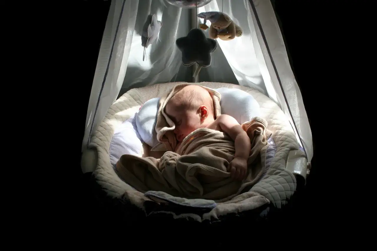 Un neonato dorme sereno nella sua culla, avvolto in una coperta. Un mobile con stelle e un orsetto pende sopra, suggerendo come far addormentare un neonato con dolcezza.