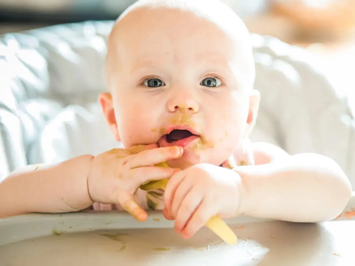 Bambino con viso sporco di pappa, intento a esplorare il cibo con le mani. Un momento di gioia e scoperta per i tagli sicuri svezzamento.