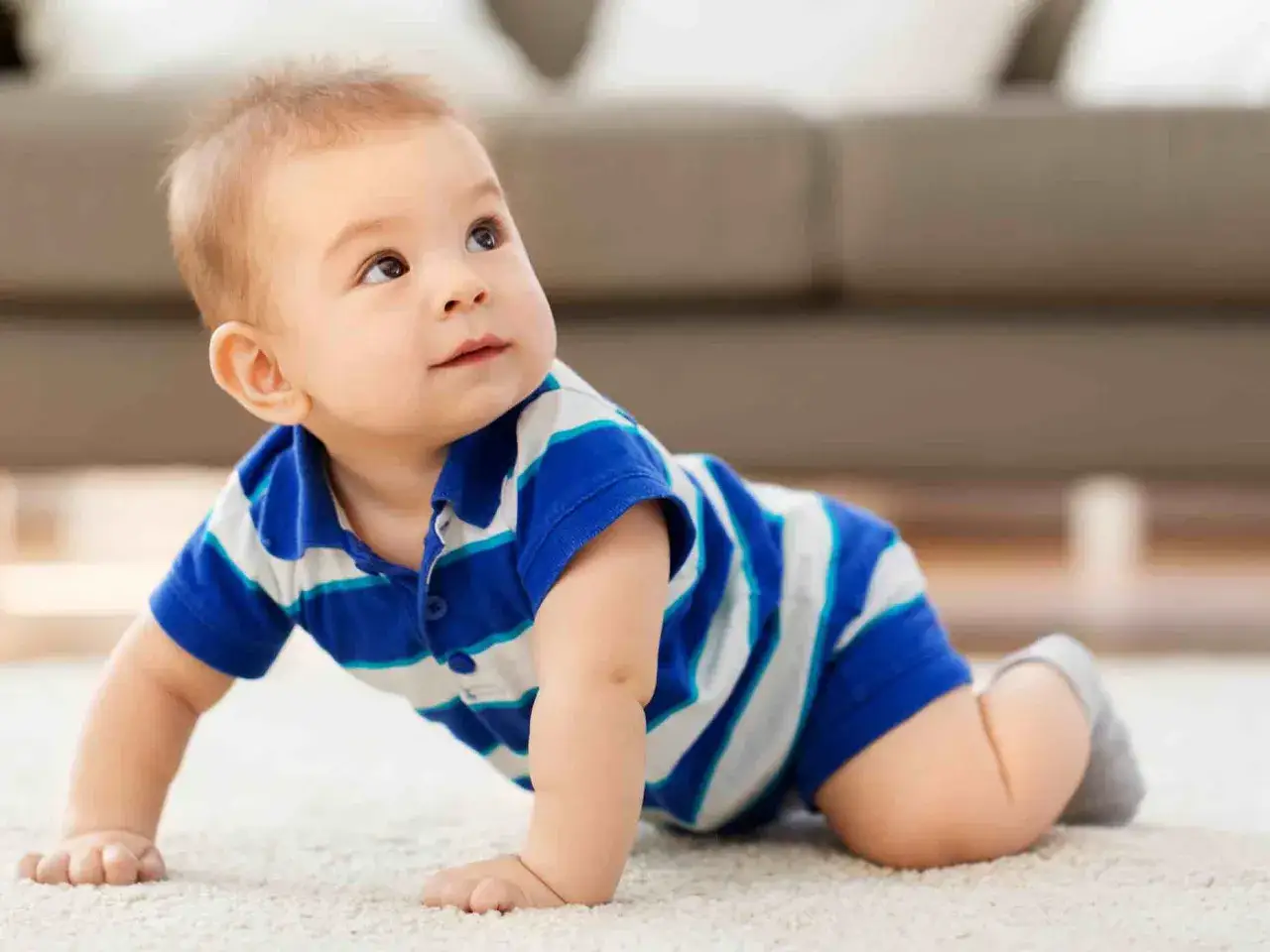 Un adorabile neonato di 8 mesi, vestito con una polo a righe blu e bianche, gattona su un tappeto chiaro, esplorando il mondo con curiosità.