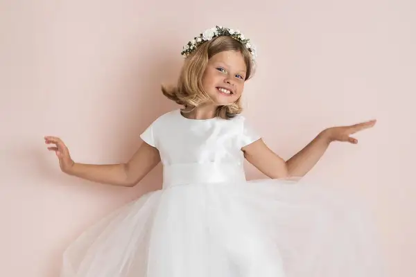 A smiling young girl in a white dress and flower crown, with arms outstretched.