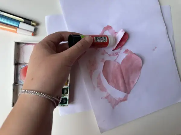 Mano che incolla un cuore di carta su un foglio, creando bellissimi disegni per la mamma. Vicino, acquerelli e pennarelli.