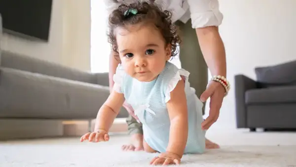 A baby in a blue striped outfit crawls on a white rug, with a person's hands gently supporting them.