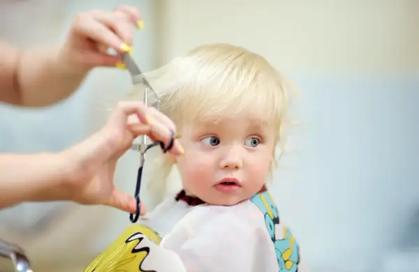 Bambino biondo con occhi azzurri riceve il suo primo taglio capelli, un momento speciale immortalato con cura.