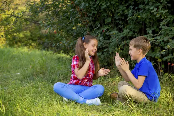 Bambini seduti sull'erba, intenti in divertenti giochi con le mani, ridono felici sotto gli alberi.