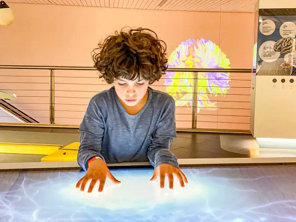 Bambino esplora un tavolo interattivo in uno dei musei per bambini, con un'installazione luminosa a forma di cervello sullo sfondo.