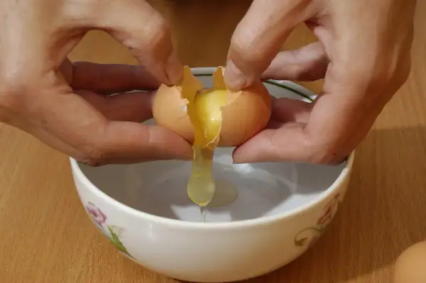 Mani che rompono un uovo per preparare uova alla coque, con il tuorlo che cola in una ciotola.
