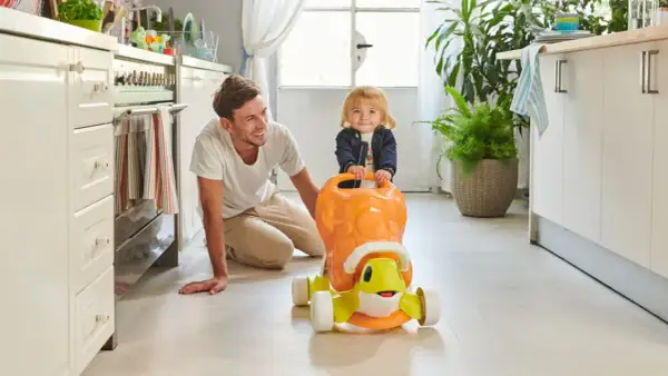 Un papà sorridente incoraggia i primi passi del suo bambino su un cavalcabile a forma di tartaruga in cucina.
