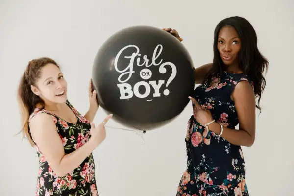 Due donne sorridono tenendo un palloncino nero con scritto "Girl or Boy?". Il **gender reveal significato** è l'attesa di una nuova vita.